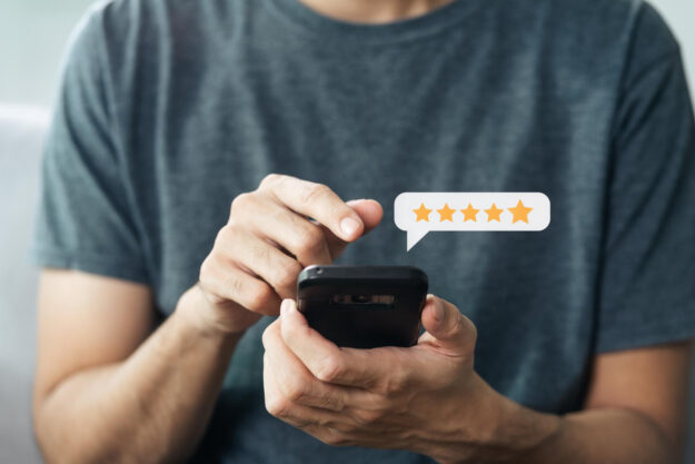 Een persoon met een grijs t-shirt aan, heeft een mobiel in zijn hand. Met zijn andere hand wijst hij naar het scherm. Boven het mobiel staat een tekstvak met vijf sterren om een ervaring achter te laten.