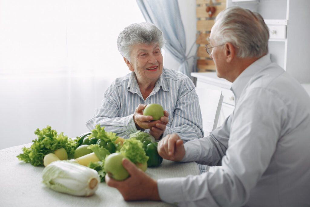 Man en vrouw met artrose zitten aan een tafel met groente en fruit die vitamine k bevatten.